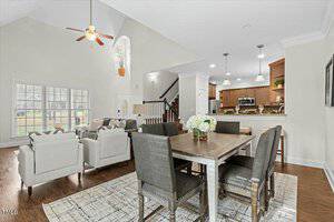 Open-concept living and dining area with high ceilings. A dining table with six chairs is in the foreground. The kitchen has stainless steel appliances. A cozy living space features sofas near a staircase. Ceiling fan and hanging lights are visible.