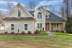 A two-story house located at 2100 Troys Trail in Franklinton features light gray siding, a dark gray roof, and stone accents around the entrance. The front boasts several windows, including an arched one above the door. A concrete pathway graces the yard, surrounded by trees.