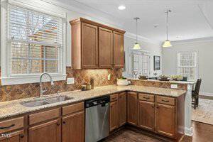 A kitchen with wooden cabinets, granite countertops, and a tiled backsplash. There is a stainless steel sink and dishwasher on the left, with two hanging pendant lights above. A dining area with windows is visible in the background.