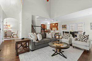 A modern living room with a gray sofa and two beige armchairs arranged around a round wooden coffee table. The space features hardwood floors, light walls, and an open view into a dining area and kitchen with brown cabinetry and pendant lights.