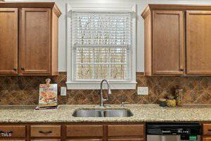 Kitchen interior featuring a granite countertop, dual sink, wooden cabinets, and a window with blinds. A cookbook stands near the left side, and a dishwasher is installed below the counter on the right.