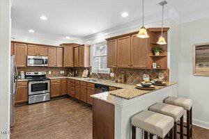 A spacious kitchen with wooden cabinets, stainless steel appliances, and a granite countertop on an island with three stools. The flooring is dark wood, and there are pendant lights hanging above the island. A small window lets in natural light.