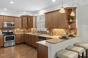 A kitchen with wooden cabinets and granite countertops. There is a stainless steel oven and microwave on the left, a dishwasher under the counter, and a double sink under a window. Three stools line a breakfast bar on the right. Pendants hang above.