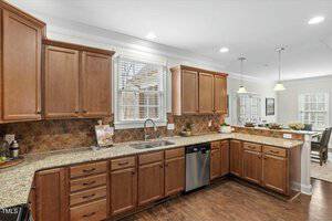 A kitchen with wooden cabinets, granite countertops, and a double sink. There is a dishwasher under the counter and a backsplash with a brown tile pattern. Pendant lights hang over the counter, and a dining area is visible in the background.