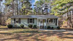 Nestled in a wooded area at 749 Fred Royster Road, Henderson, this small, single-story house features light green siding and a metal roof. A pathway meanders past several bushes to a porch with white railings, all surrounded by tall trees basking in the sun.