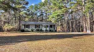 A small single-story house with a gray exterior and white trim sits in a clearing surrounded by tall trees. The lawn is sparse with brown grass, and a gravel driveway leads up to the entrance. The sky is clear with a few clouds.