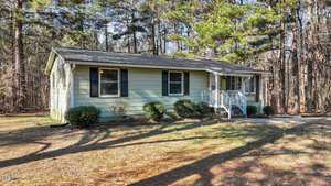 Small single-story light green house with a dark roof and white trim set amid tall trees. It has a front porch with steps, black shutters, and a surrounding yard with sparse bushes and pine needles on the ground.