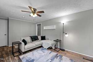 A living room with a beige sectional sofa and dark cushions, a patterned rug on wooden flooring, a floor lamp, and an overhead ceiling fan. Walls are painted light gray with a white door visible in the background.