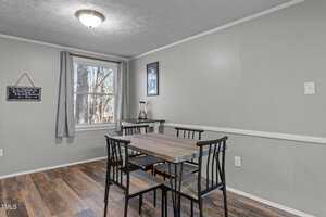 A dining area with a rectangular wooden table and four black chairs on a wood-look floor. A window with gray curtains is on the left, and a chalkboard hangs on the wall. A blender sits on a small table under a framed picture.