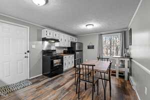 A small kitchen with white cabinets, black appliances, and a wooden dining table with four chairs. A window to the right has a view of trees outside. The floor is wood, and the walls are painted light gray. A light fixture hangs from the ceiling.