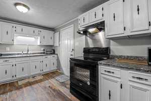 A kitchen with white cabinets and a black stove. The countertops are dark gray with a speckled pattern. There is a window above the sink on the left. A small rug lies on the wooden floor near the stove and a cutting board is on the counter.