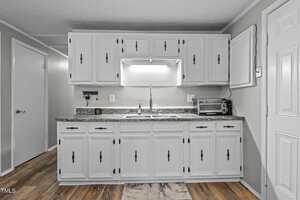 A kitchen with white cabinets and a gray countertop. There is a double sink below a bright light fixture, a small toaster oven to the right, and a soap dispenser on the countertop. The floor has a wood-like pattern.