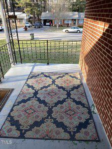 A porch with a decorative rug featuring a floral pattern in shades of pink, blue, and cream. The porch has a metal railing and a brick wall. In the background, there is a residential street with several houses and parked cars.