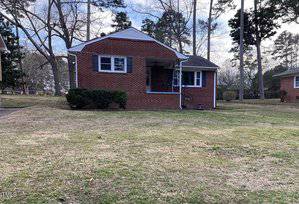 A single-story brick house with a small covered front porch and two windows visible. The front yard is grassy with a few shrubs near the house. Several tall trees stand in the background. The sky is partly cloudy.