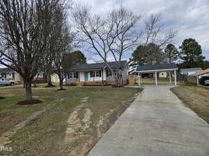 The single-story house at 912 Hillsboro Street features gray siding, a welcoming front porch, and a carport on the right. The driveway connects from the street to the carport, with bare trees in the front yard and other charming Oxford homes visible in the background.