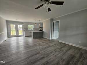 Spacious empty room with light gray walls and wooden flooring. A ceiling fan is mounted above. In the background, there's an open kitchen with a large island, white cabinets, and pendant lights. French doors on the left open to a view of the outdoors.