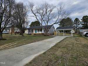 A single-story house with gray siding and a dark roof stands in a yard. The driveway leads to a carport on the right. The yard has sparse grass and a few leafless trees. A shed is visible in the background. Other houses are nearby.