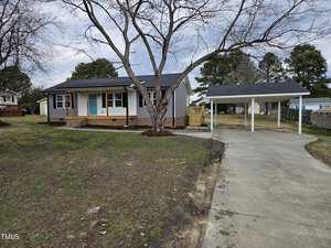 Single-story house with gray siding and a blue front door. A leafless tree stands in the front yard. The driveway leads to a carport on the right. The yard is mostly bare with patches of grass. Steps lead to a small porch at the entrance.