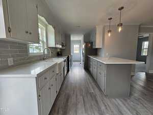 A modern kitchen with white cabinets, light gray countertops, and a gray tile backsplash. It features stainless steel appliances, including a refrigerator and stove. Pendant lights hang over a central island with seating space. Wood-look flooring.