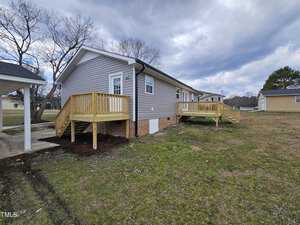 A single-story gray house with brick foundation features newly built wooden decks on the side and back. The yard is grassy, and there are a few leafless trees. The sky is cloudy with patches of blue.