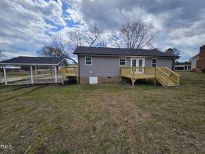 A single-story gray house with a black roof and wooden deck is situated on a grassy lawn. There is a covered carport to the left, and a chimney is visible on the right. The sky is partly cloudy.