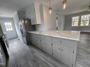 A modern kitchen with light gray cabinets and a marble countertop island. Stainless steel refrigerator is to the left, and pendant lights hang above the island. Large windows and a door provide natural light from the back wall. Floors are light wood.