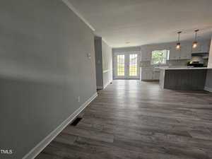A modern, empty living space with gray walls and laminate flooring leading to an open kitchen. The kitchen features white cabinets, a central island, pendant lighting, and a door with windows that opens to the outside.