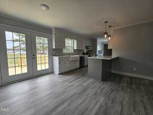 A modern kitchen features light gray hardwood floors, white cabinetry, and a center island with a counter. Pendant lights hang above the island. Large glass doors on the left lead to an outdoor area with a tree visible outside.