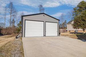 A large, gray metal garage with two closed white roll-up doors sits on a concrete driveway. The surrounding area includes grass, a few trees, and a neighboring house partly visible to the right. The sky is clear with some clouds.