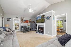 A living room with beige carpet and gray walls features two beige recliners and a large sectional sofa. The room includes a ceiling fan, a fireplace with a TV above it, and wall decor like deer mounts and an American flag. A dining area is visible through a doorway.