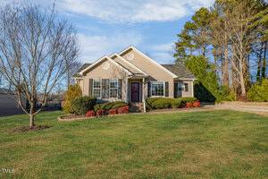 Located at 153 Bogey Lane in Henderson, this charming single-story house features beige siding and a gray shingle roof. It rests on a lawn adorned with sparse shrubs, flanked by a leafless tree to the left and tall trees in the background beneath a partly cloudy sky.