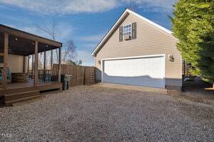 A gravel driveway leads to a beige garage with a white door. The garage is attached to a house with a covered wooden porch on the left. A ladder leans against a wooden fence. Tall trees are visible in the background under a partly cloudy sky.