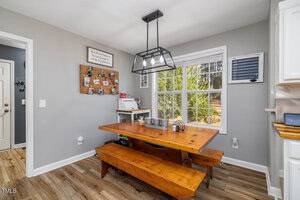 A dining area with a wooden table and benches on a brown floor. A black pendant light hangs above. A window with white trim shows outdoor greenery. A bulletin board with photos is on the gray wall, alongside a sign and wall organizer.