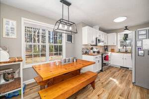 A kitchen with wooden flooring features a wooden table and benches. The room has white cabinets, a stainless steel oven, and a refrigerator. A large window provides natural light. A ceiling light hangs above the table, and a mixer is on the counter.
