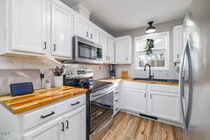 Modern kitchen with white cabinets and wood countertops. Includes a stainless steel stove, microwave, and refrigerator. A black sink is beneath a window with blinds. A small device is on the counter, and the floor is light wood.