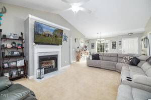 A living room with beige carpet, featuring a gray sectional sofa and a dark recliner. A fireplace with a TV above is against one wall. Shelves with pictures are to the left, and a door with windows is at the back. A ceiling fan hangs above.