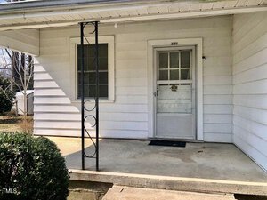 A small covered porch with a concrete floor and decorative metal support on the left. There is a window with a blind on the left side and a closed door with a glass panel and wreath on the right. A hedge is visible in front of the porch.