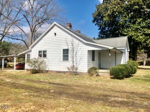 A white, single-story house with a gable roof and a small porch. There's a carport to the left, partially visible. The yard has sparse grass and a few small bushes. Trees without leaves are in the background under a clear blue sky.