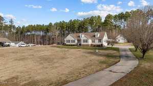 A house with a beige exterior sits on a large plot of land, featuring a long driveway leading to the property. The surrounding area is grassy with a few trees and a wooded area in the background. Several vehicles are parked near the house.