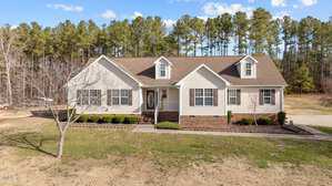A single-story house at 1031 Franklin Lane is pictured, showcasing beige siding and a brown shingled roof. It features white trim, a central front door with sidelights, and small shrubs along the front. The home in Henderson is set against tall pine trees and a blue sky.