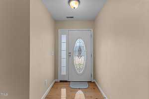 A hallway with light beige walls and wooden flooring leads to a white front door with an oval, decorative glass panel. A ceiling light fixture illuminates the space, and a small rectangular rug lies in front of the door.