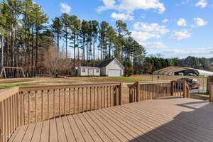 A wooden deck overlooks a spacious backyard with a shed and a garage. Tall trees line the perimeter. A swing set is on the left, and a carport shelters a red vehicle on the right. The sky is clear with scattered clouds.