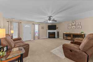 Spacious living room with beige carpet and light brown walls. Features brown reclining sofas facing a fireplace with a TV above it. A ceiling fan hangs above, and three windows and a door provide natural light. Decorative items and wall art adorn the space.
