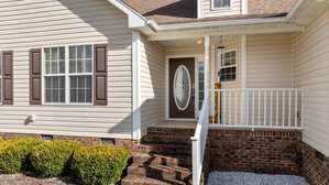 The image shows the entrance of a house with beige siding, brown shutters, and a brown door with an oval window. There are brick steps leading up to a small porch with white railing. Shrubs line the base of the house, and gravel is visible beside the steps.
