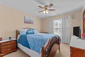 A bedroom with a wooden bed frame and blue bedding. There's a wooden nightstand with a lamp on the left and a dresser with a TV on the right. A ceiling fan is above, and a window with white curtains lets in natural light. A painting hangs on the wall.