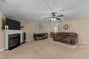 A living room with beige walls and carpet features a dark wood fireplace with a TV on top. There is a brown leather sofa, a sideboard with picture frames above it, and a ceiling fan with lights. A small dining area is visible in the background.