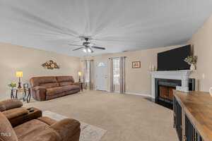 A spacious living room with beige walls and carpet features a brown reclining sofa and chair. A fireplace with a TV above it is on the right. There's a ceiling fan with lights, two small windows, and a white front door in the background.