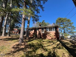 Set on a slight hill at 1220 Roanoke Avenue, this brick house with a small porch is enveloped by tall pine trees. A wooden staircase leads to the entrance under a clear blue sky, hinting at sunshine. The neighboring Henderson home peeks through the foliage.