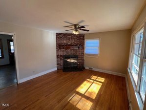 A living room with hardwood floors and a brick fireplace. There's a ceiling fan with lights above, a window with blinds to the right, and sunlight casting shadows on the floor. An open doorway leads to the adjacent room.