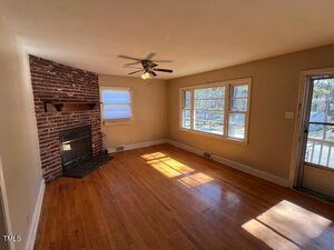 Living room with hardwood floors, a ceiling fan, and a brick fireplace on the left wall. A pair of windows and an exterior door allow natural light in from the right side. Walls are painted light beige.